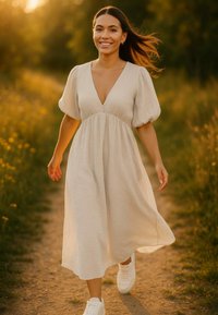 Een vrouw in een beige, knie-lange jurk met een diepe V-hals en pofmouwen, gecombineerd met witte sneakers, die langs een graspad loopt.