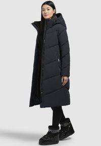 Lang sort pufferjakke med hætte, der har et quiltet design, lynlås og knaplukninger samt sidelommer. Sammen med sorte, isolerede støvler.