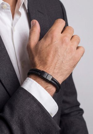 Man's hand adjusting black braided leather bracelet with metal clasp, wearing dark suit jacket and white shirt cuff.