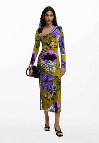 Langärmliges, figurbetontes Kleid in Olivgrün mit großen lila und weißen Blumenmustern. Kombiniert mit einer schwarzen Handtasche und Sandalen mit Absatz.