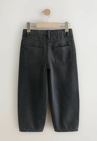 Pantaloni in denim neri con una vestibilità rilassata, appesi a un appendiabiti in legno. La parte posteriore presenta due tasche e cuciture visibili.