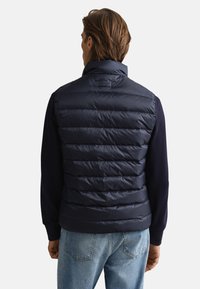 Marinblå vadderad väst med horisontell sömnad, hög krage och dragkedja, bärs över en mörk tröja, med en slät, glänsande yta.