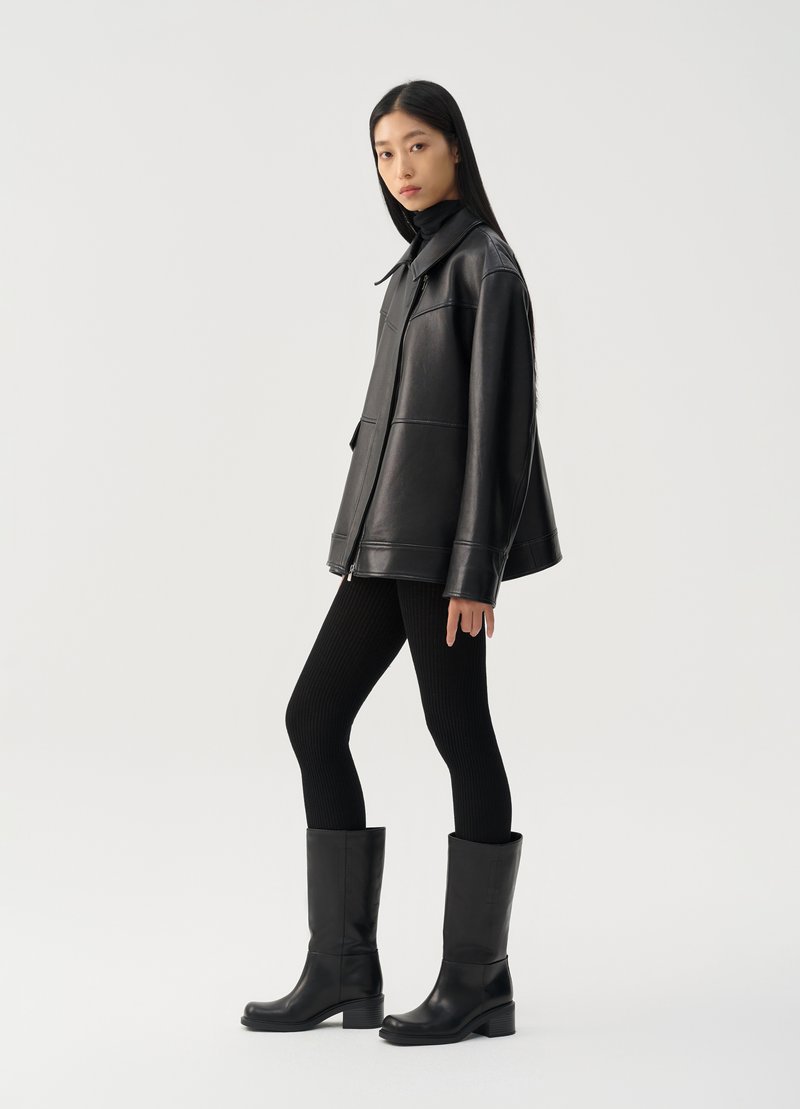 Giacca di pelle nera con colletto, abbinata a leggings neri a costine e stivali neri al ginocchio. Texture liscia e design minimalista.