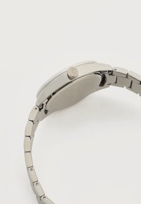 Ασημένιο μεταλλικό ρολόι χειρός, εμφανιζόμενο από το πλάι, που δείχνει το κορόνα και το συνδεδεμένο βραχιόλι σε ένα απλό λευκό φόντο.