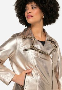 Metallisch goldene Bikerjacke mit Nietenakzenten, Reißverschlusstaschen, Halskragen mit Kettendetails und taillierter Passform. Glatte Textur und glänzende Oberfläche.