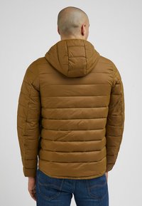 Brązowa kurtka puchowa z kapturem, z poziomymi przeszyciami, gładką fakturą i dopasowanym fasonem, noszona z niebieskimi dżinsami.