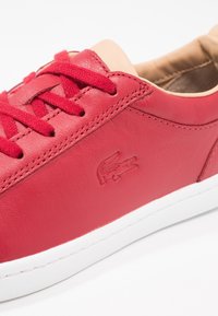 Röda lädersneakers med röda snören, beige vadderad krage, vit sula och präglad krokodillogga på sidan.