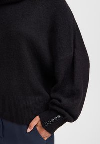 Pull en tricot noir avec une silhouette ample, un ourlet à côtes et des détails de boutons sur les poignets. Texture douce avec une coupe décontractée.