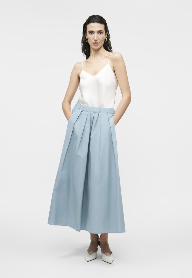Femme aux longs cheveux noirs portant un caraco blanc, un pantalon large bleu clair, des talons blancs pointus et des boucles d'oreilles épaisses dorées, debout les mains dans les poches.