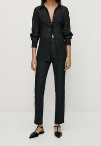 Zwarte transparante blouse met lange mouwen, gecombineerd met zwarte high-waisted jeans en puntige ballerina's, wat een monochromatische look creëert.