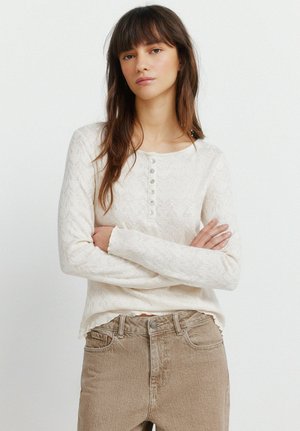 Giovane donna con capelli castani che indossa un maglione crema a maniche lunghe con bottoni e jeans marrone chiaro, con le braccia incrociate, sfondo neutro.