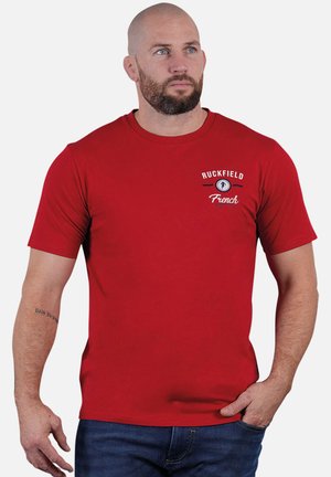Homme avec une barbe et un tatouage sur le bras portant un T-shirt rouge à manches courtes avec le logo "Ruckfield French Football", habillé en jean bleu.