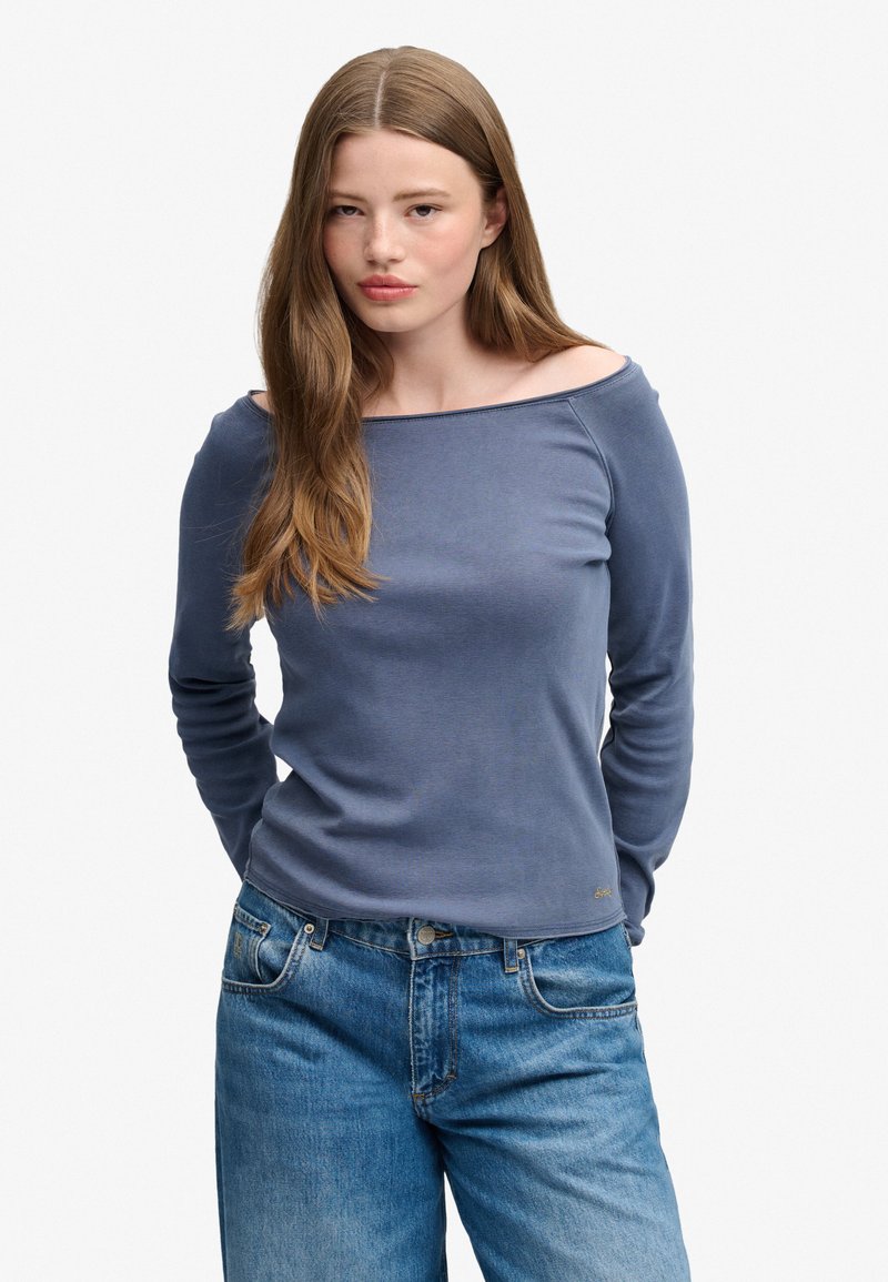Joven mujer con cabello largo y castaño, que lleva una camiseta azul de manga larga con los hombros descubiertos y vaqueros azules de corte relajado, de pie frente a un fondo liso.