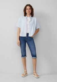 Jasnoniebieska koszula z krótkim rękawem zapinana na guziki, noszona na białej koszulce, w połączeniu z niebieskimi dżinsowymi spodniami w stylu capri i beżowymi klapkami.