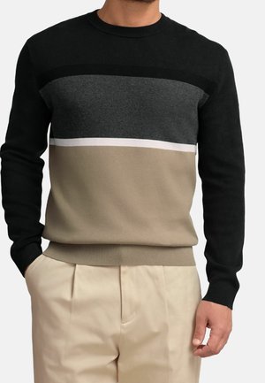 Homme portant un pull en maille à manches longues avec des blocs de couleurs horizontaux noirs, gris foncé, blancs et beiges, assorti à un pantalon beige.