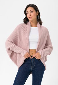 Helles rosa gestricktes Cardigan mit geripptem Muster, offener Vorderseite und weiten Ärmeln. Kombiniert mit einem weißen Crop-Top und dunkelblauen Jeans.