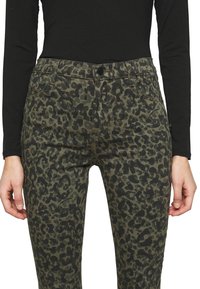 Pantalones con estampado de leopardo en verde oliva y manchas negras, con cierre frontal de botón, pasadores para cinturón y bolsillos laterales. Textura de tela suave.