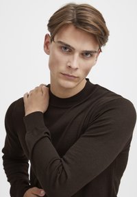 Maglione marrone lavorato a maglia con scollatura rotonda, polsini a coste, texture liscia e design aderente, indossato da un modello con capelli corti.