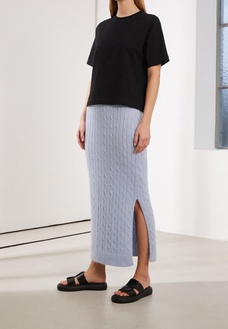 Femme portant une chemise noire à manches courtes, une jupe midi bleu clair en maille torsadée avec une fente sur le côté, et des sandales plates noires, debout à l'intérieur.