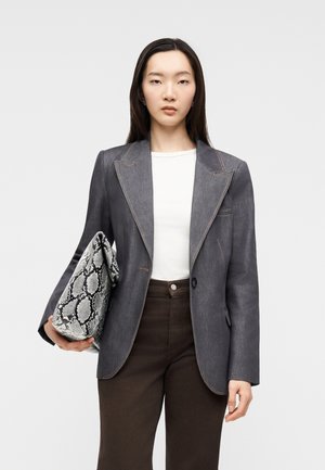 Kvinde med langt mørkt hår iført en grå blazer, hvid top og mørke bukser, der holder en stor clutch-taske med slangeskindsprint.