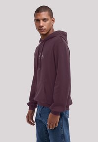 Bordeauxroter Hoodie aus weichem, dickem Stoff mit einer Vordertasche, verstellbarem Kordelzug und gerippten Bündchen. Kombiniert mit blauen Jeans.