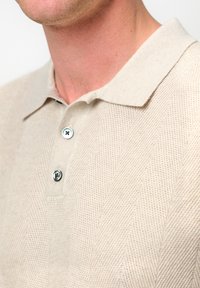 Beige stickad polo-t-shirt med ett sicksackmönster, treknapplåsning och mjuk textur. Kragen ligger platt mot nacken.