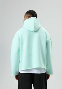 Sweat à capuche oversize vert menthe avec une grande capuche, des épaules basses et un tissu texturé. Superposé sur une chemise blanche et associé à un pantalon foncé.