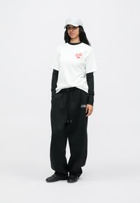 Γυναίκα με λευκό καπέλο μπέιζμπολ, λευκή μπλούζα με ροζ χείλη, μαύρη μακρυμάνικη μπλούζα από κάτω, μαύρο φαρδύ παντελόνι και μαύρα παπούτσια, σε όρθια θέση.