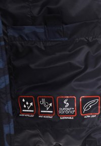 Tygetikett på mörkt material som visar ikoner och text för vattenavvisande, vindtålig, hållbar och ultralätt funktioner.