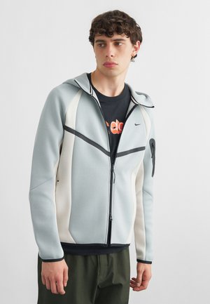 Mladý muž oblečený v svetlosivej a bielej mikine na zips cez čierne tričko s grafikou, stojaci pred jednofarebným pozadím, pozerajúci sa doľava.