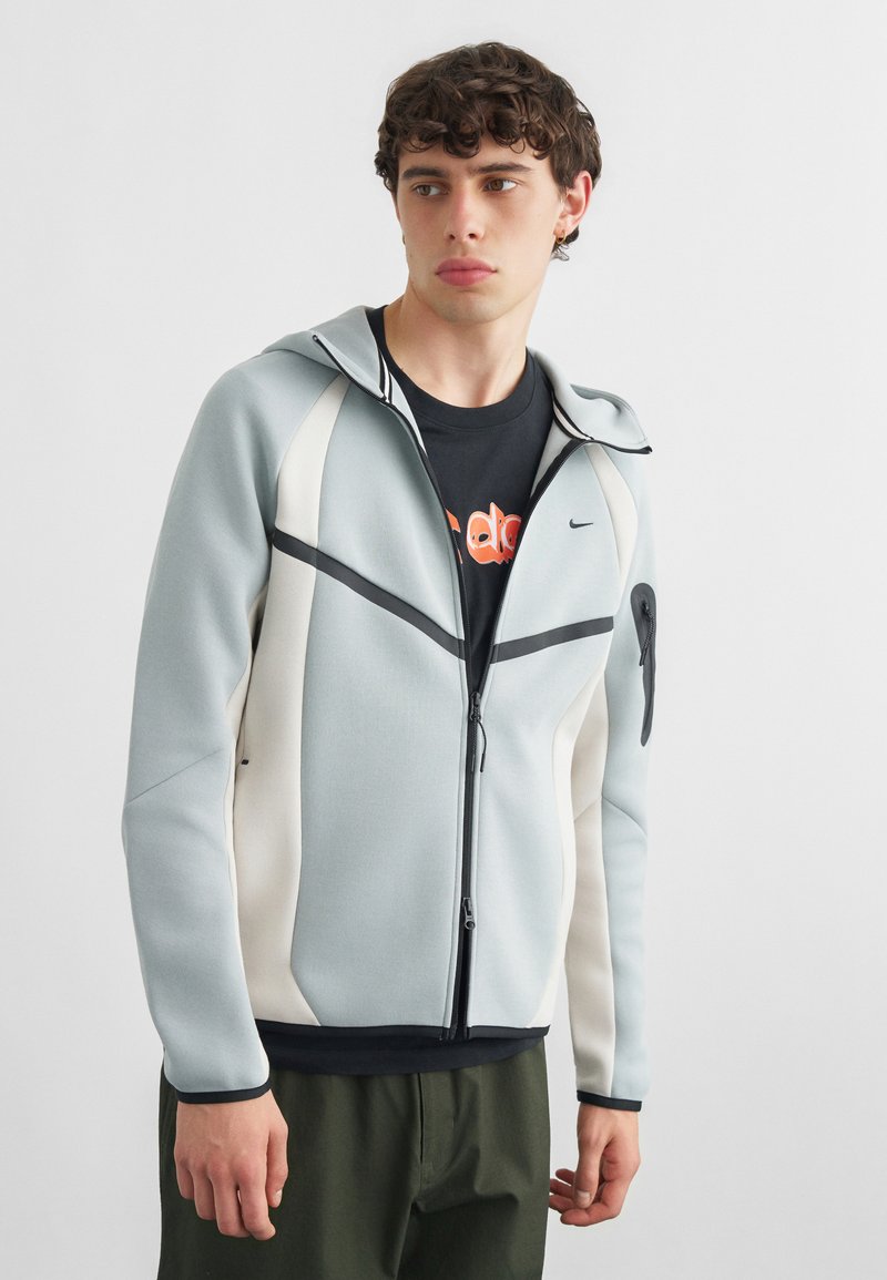 Junger Mann trägt einen hellgrauen und weißen Kapuzenpullover mit Reißverschluss über einem schwarzen T-Shirt mit Grafik, steht vor einem einfarbigen Hintergrund und schaut nach links.