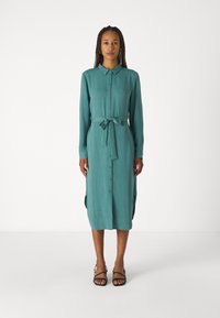 Vestito azzurro bottonato con maniche lunghe e cintura legata in vita. Realizzato in tessuto morbido, presenta spacchi laterali e una silhouette dritta. Sandali neri.