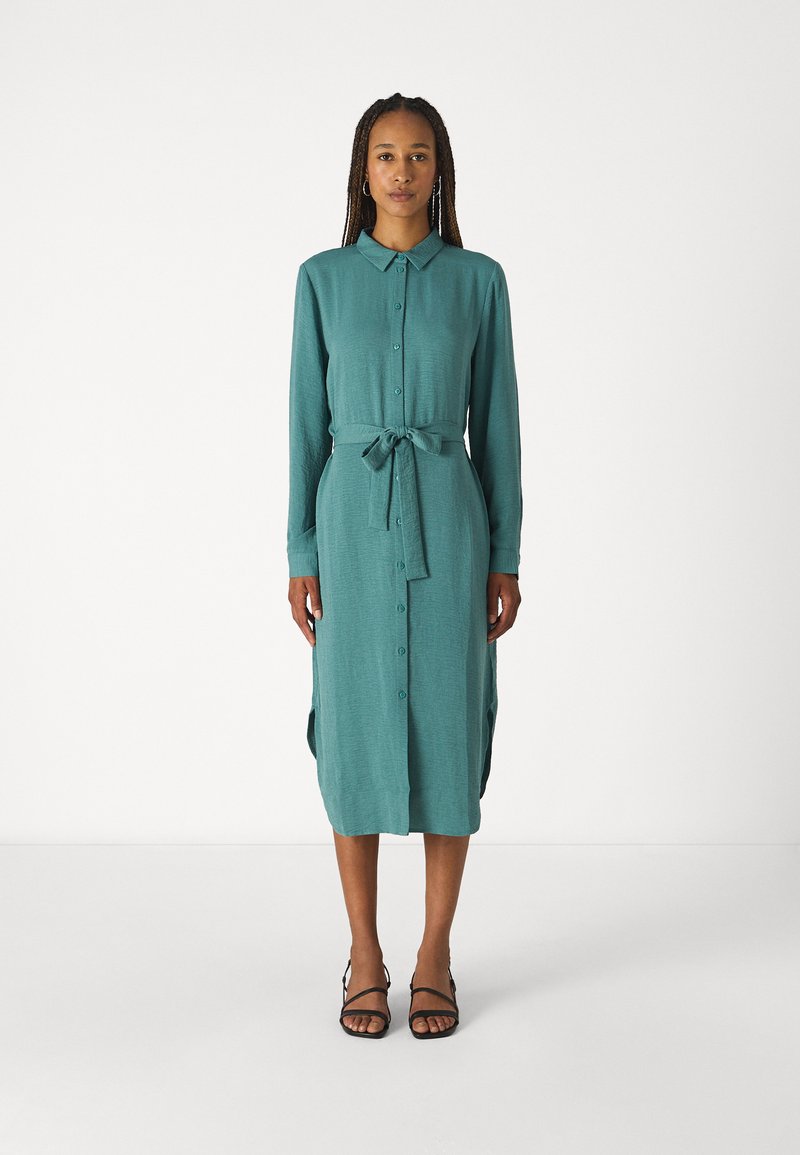 Vestito azzurro bottonato con maniche lunghe e cintura legata in vita. Realizzato in tessuto morbido, presenta spacchi laterali e una silhouette dritta. Sandali neri.