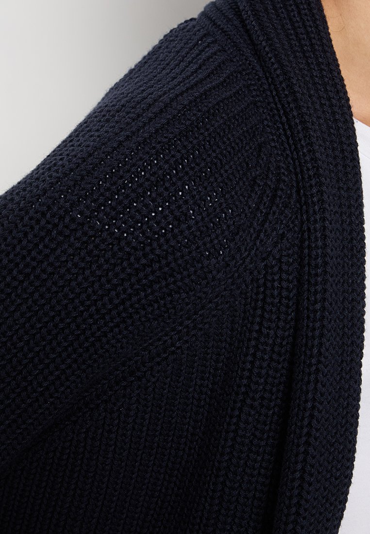 Marineblaue gestrickte Strickjacke mit gerippter Textur, offenem Frontdesign und subtilen Lochmustern im Schulterbereich. Weiches, leichtes Material.