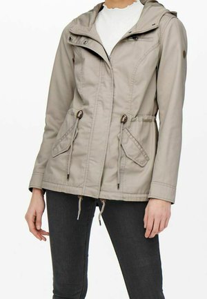 Beige jas met kap en rits, verstelbare taillekoorden, klepzakken, gedragen over een wit shirt en gecombineerd met donkere spijkerbroek.