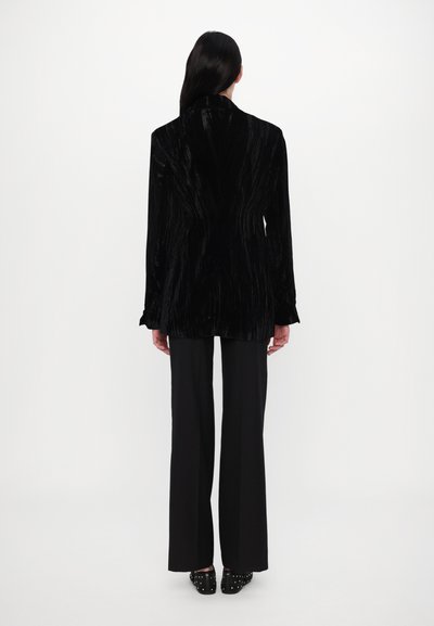 Schwarzer Samt-Blazer mit vertikalen Streifen, leicht tailliert, langen Ärmeln und Reverskragen, kombiniert mit schwarzen, weiten Hosen.