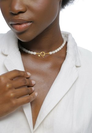 Weißes Perlen-Choker mit goldenen Hardware, ausgestattet mit einem dekorativen Verschluss und kleinen Akzenten, um den Hals getragen mit einem leichten Stoffhemd.
