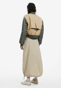 Beige and green bomber jacket with a cropped back, paired with a long beige skirt. Footwear is light-colored and minimalistic.
