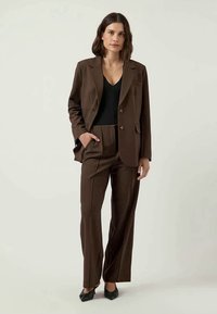 Blazer castanho sob medida e calças de cintura alta combinando, apresentando uma textura suave e uma silhueta estrutural, acompanhados de uma blusa preta.
