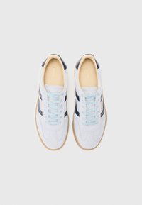 Vita mockasinsneakers med marinblå accenter, ljusblå snören och en brun gummisula. Har en rund tå och sydda detaljer på ovandelen.