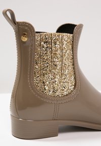 Botín impermeable marrón con un panel elástico dorado brillante, lados texturizados y un botón dorado en el borde superior.