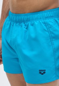Calções de banho turquesa feitos de tecido liso e leve, com cintura elástica e um pequeno logótipo preto "arena" na lateral.