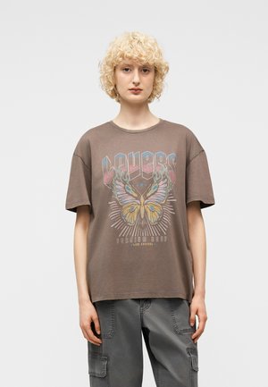 Persona con capelli biondi ricci che indossa una maglietta marrone oversize con grafica di farfalle e testo, abbinata a pantaloni cargo grigio scuro.