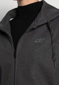 Sudadera gris con cremallera y acabado texturizado, que presenta un logo sutil y cordones con acentos rojos. Tiene un ajuste cómodo y relajado.