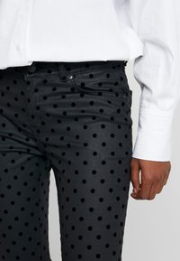 Pantalones negros con puntos de terciopelo, combinados con una camisa blanca de manga larga, mostrando la parte inferior del torso y la mano derecha de una persona.