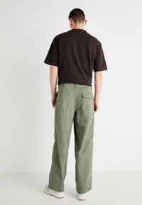 Back view of a person wearing a dark brown T-shirt and loose-fitting, light green cargo pants with large back pockets and button accents.