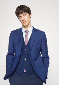 Costum albastru din trei piese, cu un sacou structurat, vestă și pantaloni adaptați. Include o cămașă de zi albastră deschisă și o cravată cu dungi.