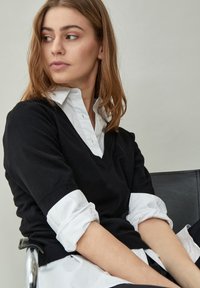 Čierny pletený sveter s V-výstrihom, nosený cez bielu košeľu s gombíkmi. Má manžetové rukávy a jemnú, hladkú textúru.