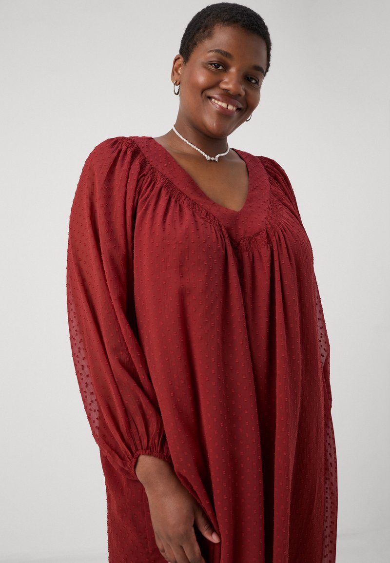 Rotes Kleid mit V-Ausschnitt, Puffärmeln und strukturiertem Punkte-Muster. Hergestellt aus leichtem Stoff, mit gerafften Details an der Brust.