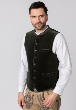 Mann trägt traditionelle bayerische Tracht mit dunkelgrüner Weste über weißem Hemd und beigen bestickten Lederhosen vor grauem Hintergrund.